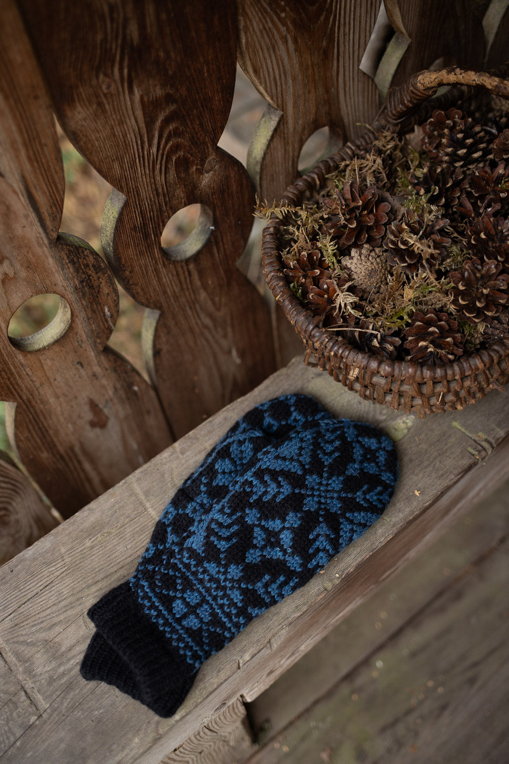 A single knit mitten in dark navy and bright blue lies on a rustic wooden surface next to a woven basket filled with pinecones and dried moss, set against a wooden fence.