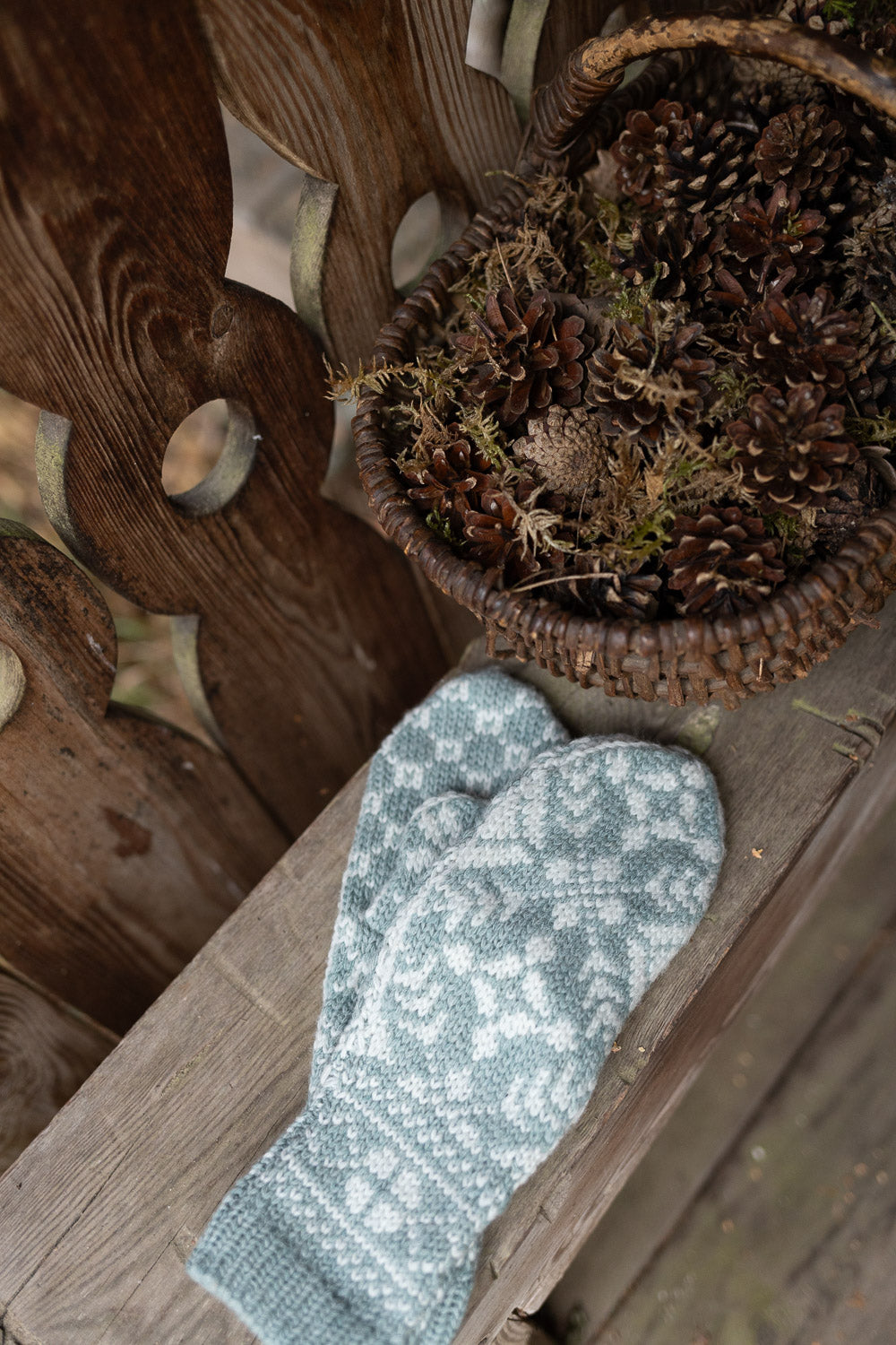A pair of knit mittens in a pale blue-gray and white pattern, resting on a rustic wooden surface next to a woven basket filled with pinecones.