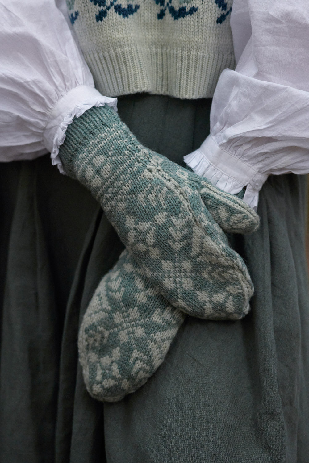A close-up of a person wearing patterned knit mittens over a dark green dress and white ruffled sleeves, with part of a knitted sweater visible at the top.