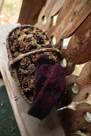 A pair of knit winter mittens in a dark red and black pattern, draped over a rustic woven basket filled with pinecones on a wooden bench.