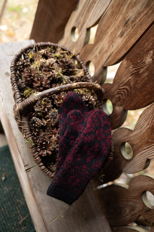 A pair of knit winter mittens in a dark red and black pattern, draped over a rustic woven basket filled with pinecones on a wooden bench.