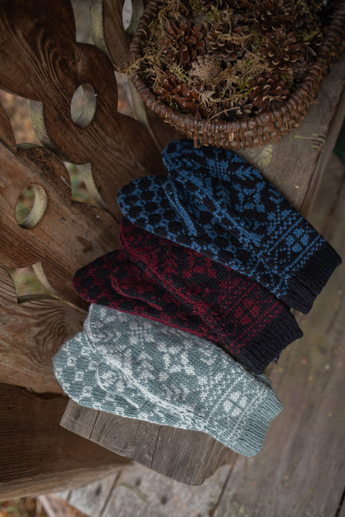 Four knit winter mittens with traditional Fair Isle patterns are arranged diagonally on a rustic wooden bench. The mittens come in four colors: light gray, dark red, navy blue, and medium blue.