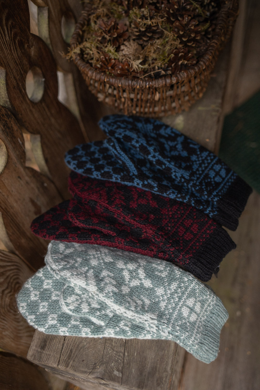 Four knit mittens with traditional Nordic patterns are stacked diagonally on a rustic wooden surface. The mittens come in four colors: light gray and white, dark red and black, blue and black, and a dark navy and black.