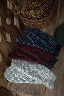 Four knit mittens with traditional Nordic patterns are stacked diagonally on a rustic wooden surface. The mittens come in four colors: light gray and white, dark red and black, blue and black, and a dark navy and black.