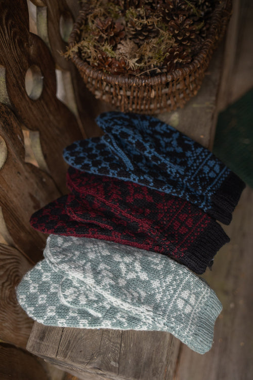 Four knit mittens with traditional Nordic patterns are stacked diagonally on a rustic wooden surface. The mittens come in four colors: light gray and white, dark red and black, blue and black, and a dark navy and black.