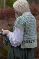 A person outdoors wearing a cream knit sweater vest with a blue and green Nordic snowflake pattern over a white blouse and dark skirt, holding a matching knit item in one hand.
