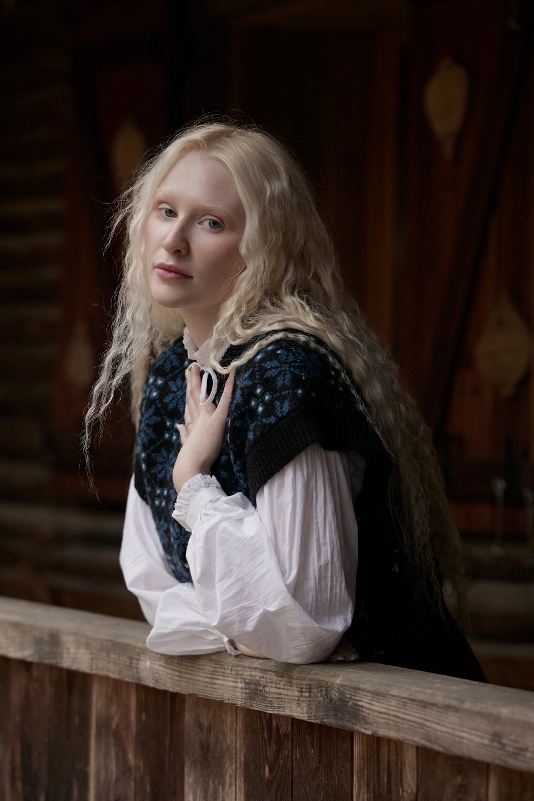 A woman with long blonde hair wearing a white blouse and a dark blue patterned vest leans on a wooden railing in a dimly lit indoor setting.