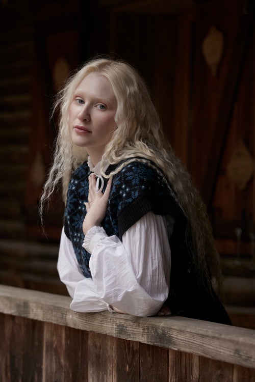 A woman with long blonde hair wearing a white blouse and a dark blue patterned vest leans on a wooden railing in a dimly lit indoor setting.