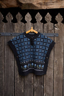 A cropped knit sweater vest with a blue and black Nordic snowflake pattern, hanging on a wooden hanger against a rustic dark wood plank wall.