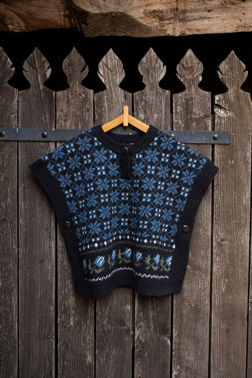 A cropped knit sweater vest with a blue and black Nordic snowflake pattern, hanging on a wooden hanger against a rustic dark wood plank wall.