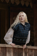 A woman with long, wavy blonde hair wearing a dark navy blue patterned knit sweater vest over a white long-sleeve blouse, leaning on a wooden railing in front of a rustic log cabin background.