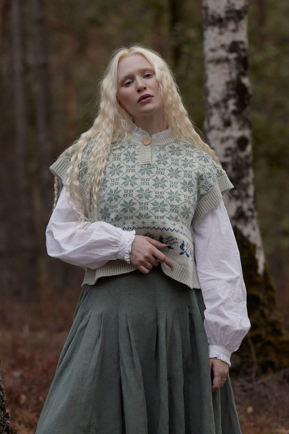 A woman with long blonde hair stands in a forest wearing a cream and sage green patterned knit vest over a white blouse and a dark green skirt.