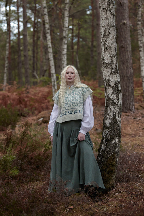 A woman with long blonde hair stands in a forest wearing a medieval-style outfit consisting of a white long-sleeve shirt, a green patterned vest, and a long green skirt, posing next to a birch tree.