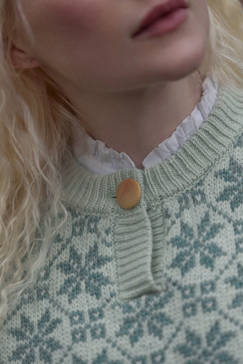 Close-up of a model wearing a light green and white patterned knit sweater with a ruffled white collar and a single wooden button.