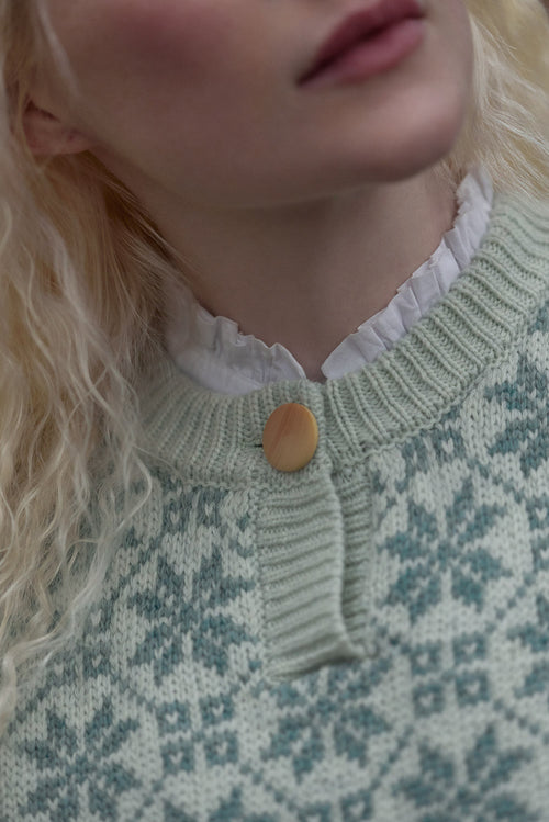 Close-up of a model wearing a light green and white patterned knit sweater with a ruffled white collar and a single wooden button.