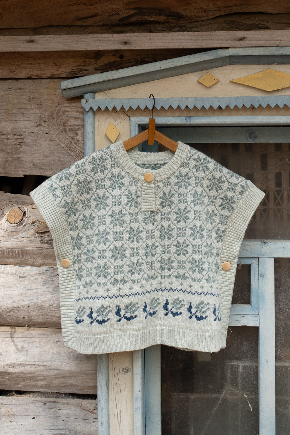 A cream-colored knit sweater vest with a gray and navy blue Nordic snowflake pattern, hanging on a wooden hanger against a rustic wooden wall and window frame.