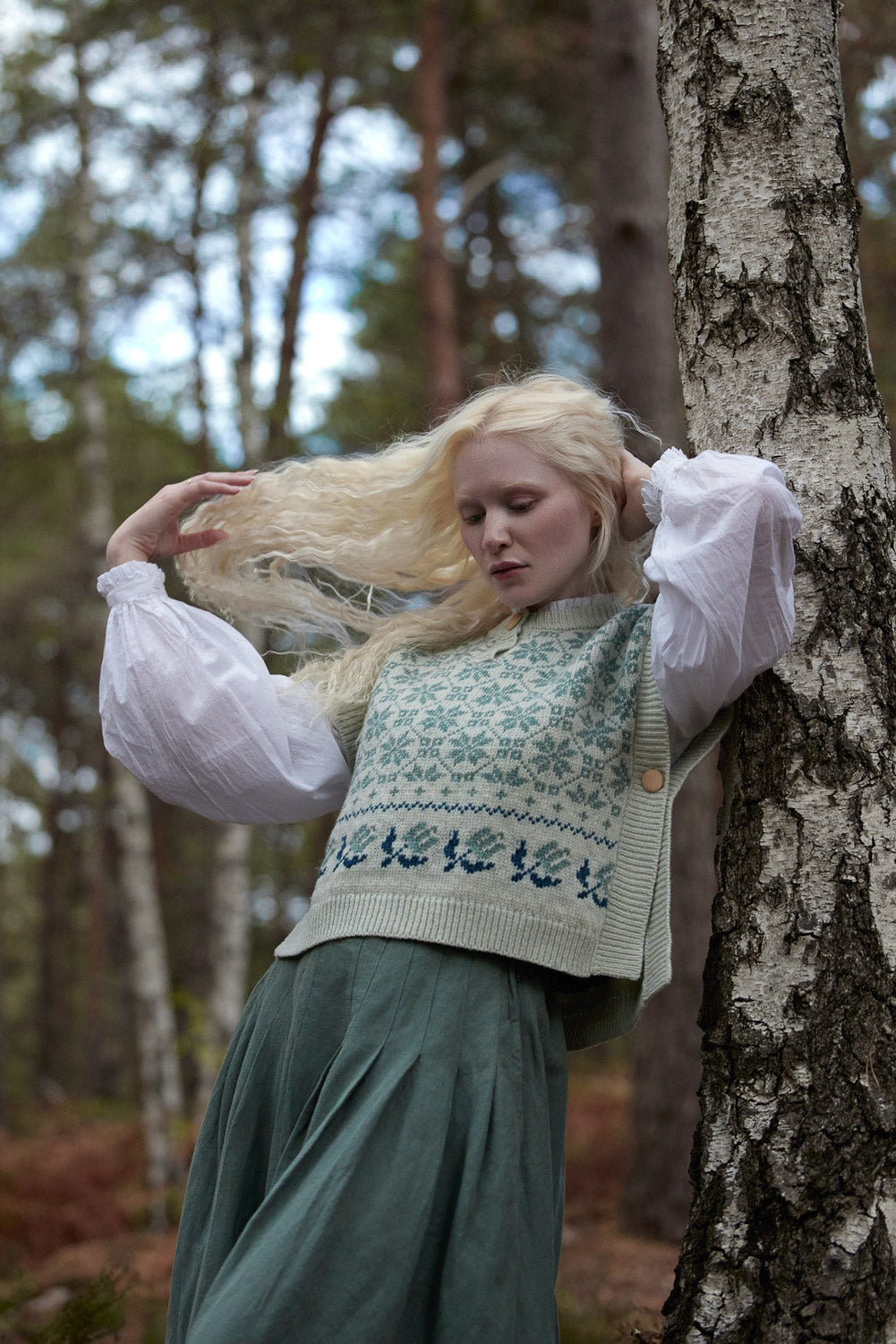 A woman with long blonde hair stands in a forest beside a birch tree, wearing a cream knit sweater vest with blue Nordic patterns over a white puff-sleeve blouse and a dark green skirt.