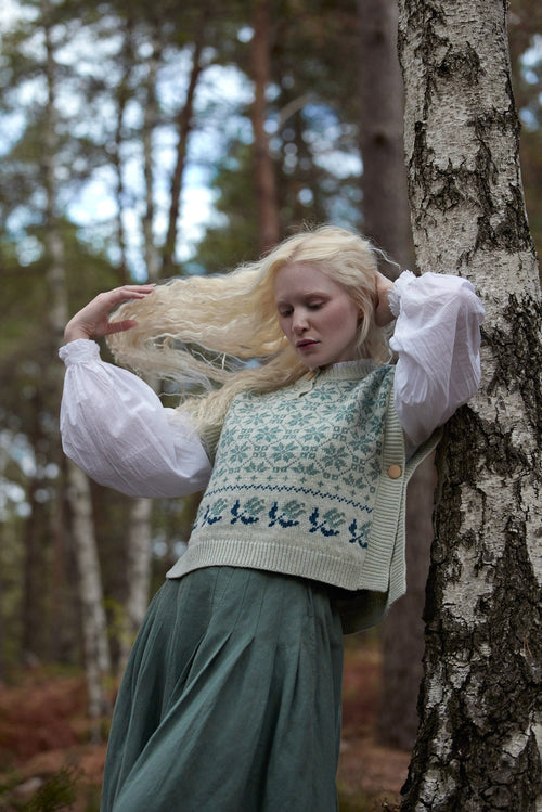 A woman with long blonde hair stands in a forest beside a birch tree, wearing a cream knit sweater vest with blue Nordic patterns over a white puff-sleeve blouse and a dark green skirt.
