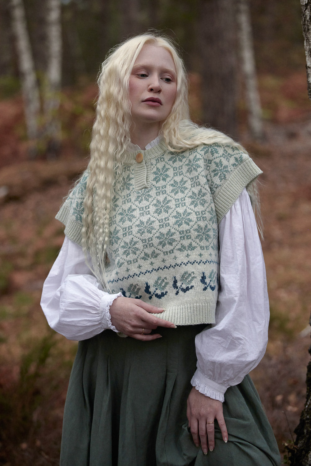 A woman with long blonde hair stands in a forest wearing a cream knit vest with a green and blue Nordic snowflake pattern over a white blouse and a dark green skirt.