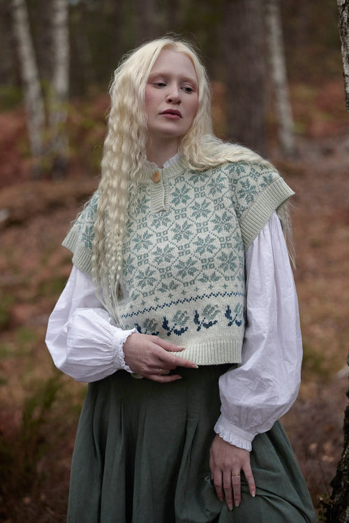 A woman with long blonde hair stands in a forest wearing a cream knit vest with a green and blue Nordic snowflake pattern over a white blouse and a dark green skirt.