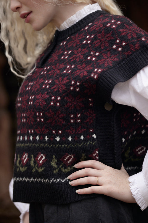 A close-up of a woman wearing a dark navy knit sweater vest with a red, white, and green Nordic/Fair Isle pattern over a white ruffled shirt.