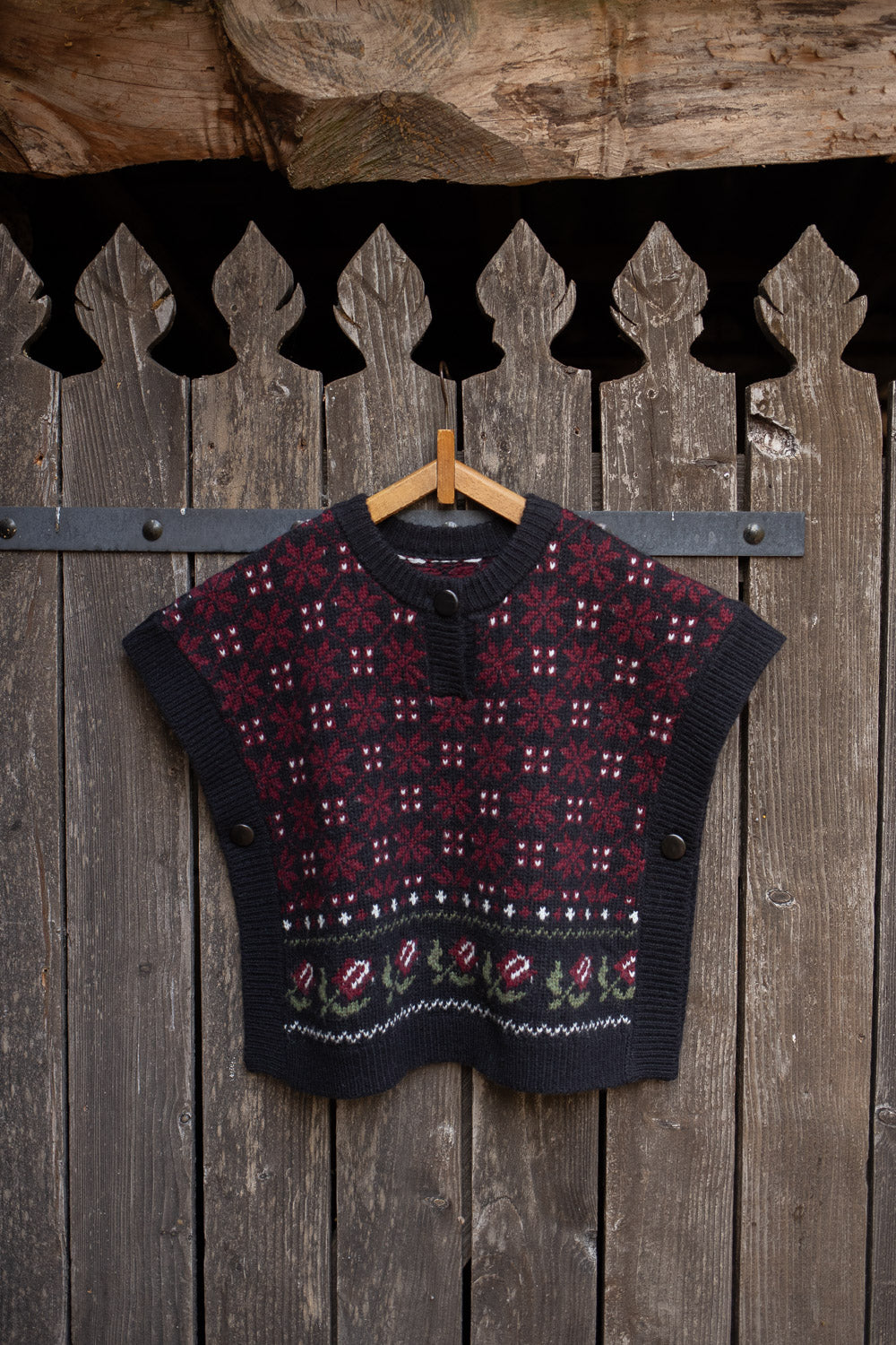A cropped knit sweater vest with a dark navy base and red, white, and green Nordic/Fair Isle patterns, hanging on a wooden hanger against a rustic dark wood plank wall.