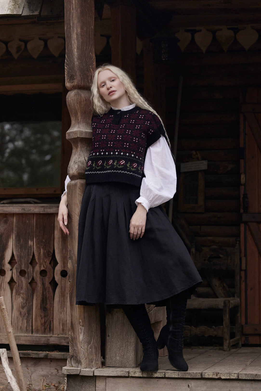 A woman stands on rustic wooden porch steps wearing a dark embroidered knit sweater vest over a white long-sleeve blouse, a dark pleated skirt, and black boots.