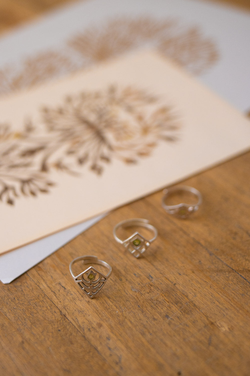 Three silver-toned rings with geometric and floral engraved designs resting on a wooden surface, with a blurred card featuring matching floral patterns in the background.