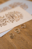 Three silver-toned rings with geometric and floral engraved designs resting on a wooden surface, with a blurred card featuring matching floral patterns in the background.