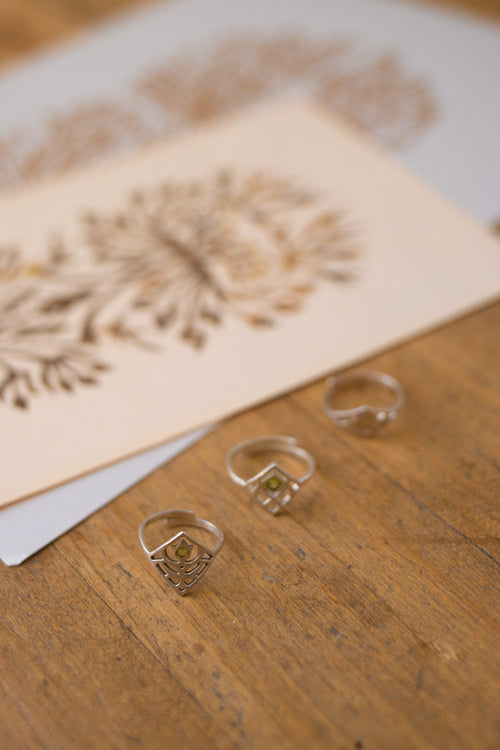 Three silver-toned rings with geometric and floral engraved designs resting on a wooden surface, with a blurred card featuring matching floral patterns in the background.