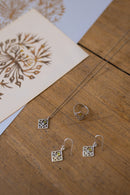 Silver jewelry set featuring a necklace with a square filigree pendant, a matching ring, and drop earrings, displayed on a wooden surface next to a decorative card.