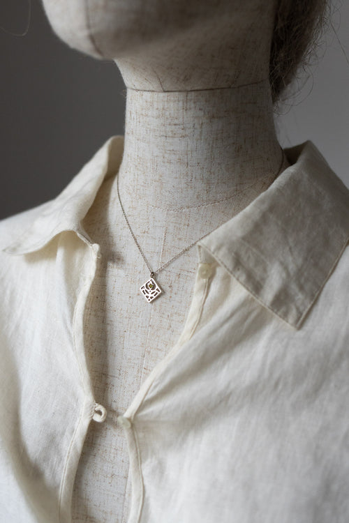 A delicate silver geometric pendant necklace displayed on a linen mannequin torso wearing a cream-colored linen button-up shirt.