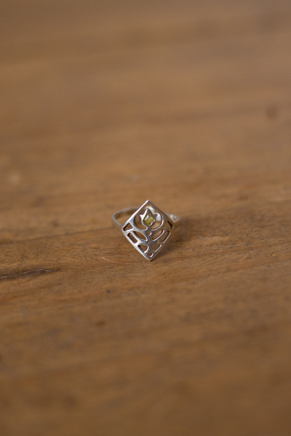 A silver ring with a geometric, openwork diamond-shaped setting and a small yellow gemstone in the center, resting on a wooden surface.