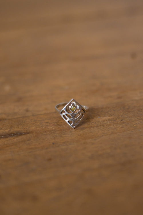 A silver ring with a geometric, openwork diamond-shaped setting and a small yellow gemstone in the center, resting on a wooden surface.
