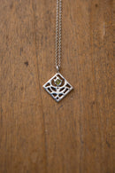 A silver geometric diamond pendant necklace with a small green gemstone in the center, hanging on a wooden surface.