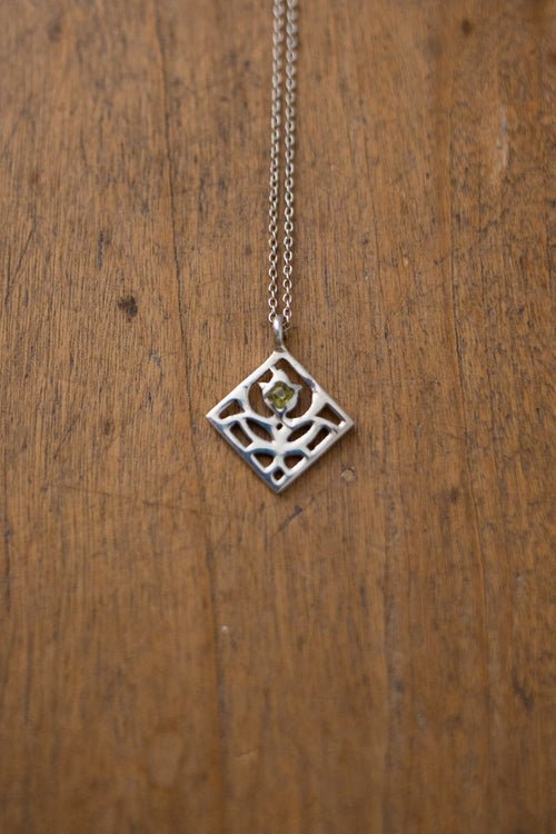A silver geometric diamond pendant necklace with a small green gemstone in the center, hanging on a wooden surface.