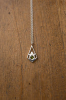 A silver geometric pendant necklace with a small green gemstone, displayed on a textured wooden surface.