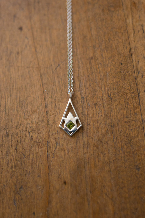 A silver geometric pendant necklace with a small green gemstone, displayed on a textured wooden surface.
