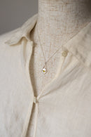 A delicate gold necklace with a small diamond-shaped pendant featuring a yellow inlay, displayed on a beige linen shirt mannequin.