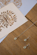 Silver geometric dangle earrings and matching necklace with small green stone accents, displayed on a wooden surface next to a printed card.