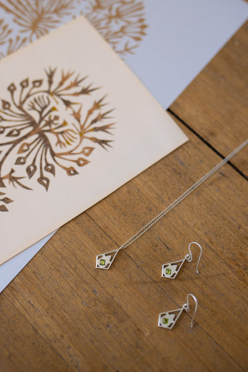 Silver geometric dangle earrings and matching necklace with small green stone accents, displayed on a wooden surface next to a printed card.