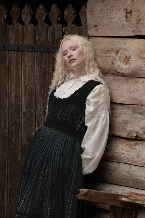 A woman with long blonde hair leans against a rustic wooden log wall, wearing a traditional-style outfit with a white blouse, a black lace-trimmed vest, and a dark pleated skirt.