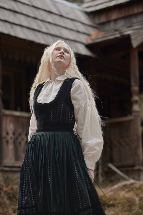 A woman with long blonde hair wearing a traditional black and white embroidered dress stands outdoors in front of a rustic wooden building.