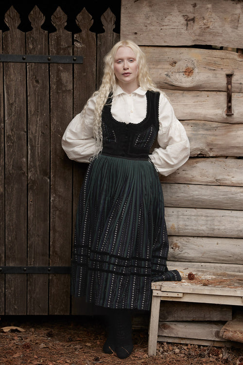 A woman stands outdoors against a rustic wooden wall and door, wearing a long-sleeved white blouse with a black embroidered bodice and a long dark green pleated skirt with white embroidered details.