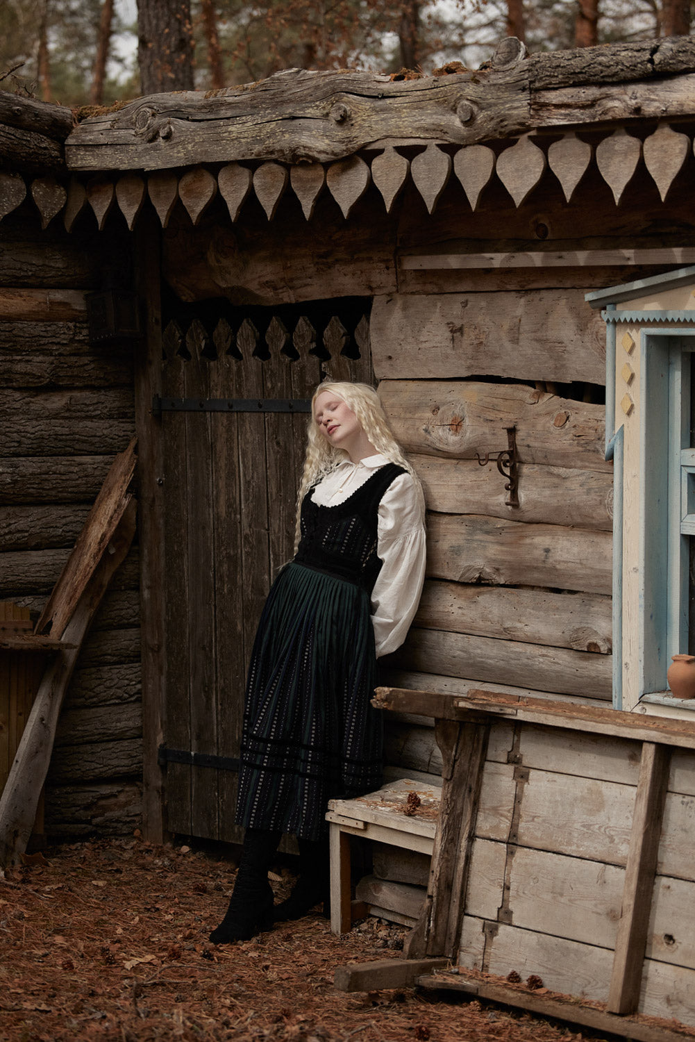 A woman with long blonde hair stands leaning against a rustic wooden cabin wall, wearing a black and white vintage-style dress with a pleated skirt and white ruffled collar. The setting is outdoors in a wooded area with fallen leaves on the ground.