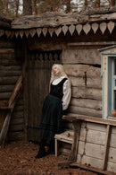 A woman with long blonde hair stands leaning against a rustic wooden cabin wall, wearing a black and white vintage-style dress with a pleated skirt and white ruffled collar. The setting is outdoors in a wooded area with fallen leaves on the ground.