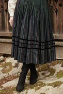 A person wearing a long pleated skirt with vertical dotted stripes in black, green, and white, paired with black lace-up ankle boots, standing outdoors on a mossy stone surface in front of a rustic wooden wall.