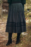 A dark green pleated midi skirt with vertical dotted stripes and horizontal banding, worn by a model in an outdoor forest setting.