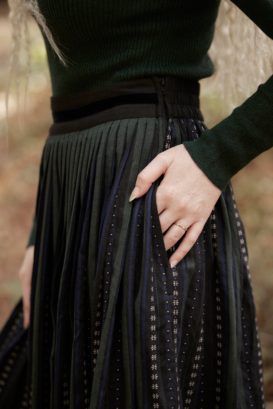 A close-up of a person wearing a dark green ribbed knit top and a long black pleated skirt with vertical dotted stripe patterns and side pockets, with one hand in the pocket against a blurred outdoor background.