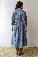 Woman wearing a crossover blue dress with floral embroideries standing on hay.